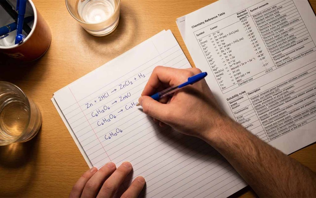 Ręka ucznia rozwiązującego zadania chemiczne na kartce, wzory i równania widoczne, obok tablica chemiczna — umieść tutaj