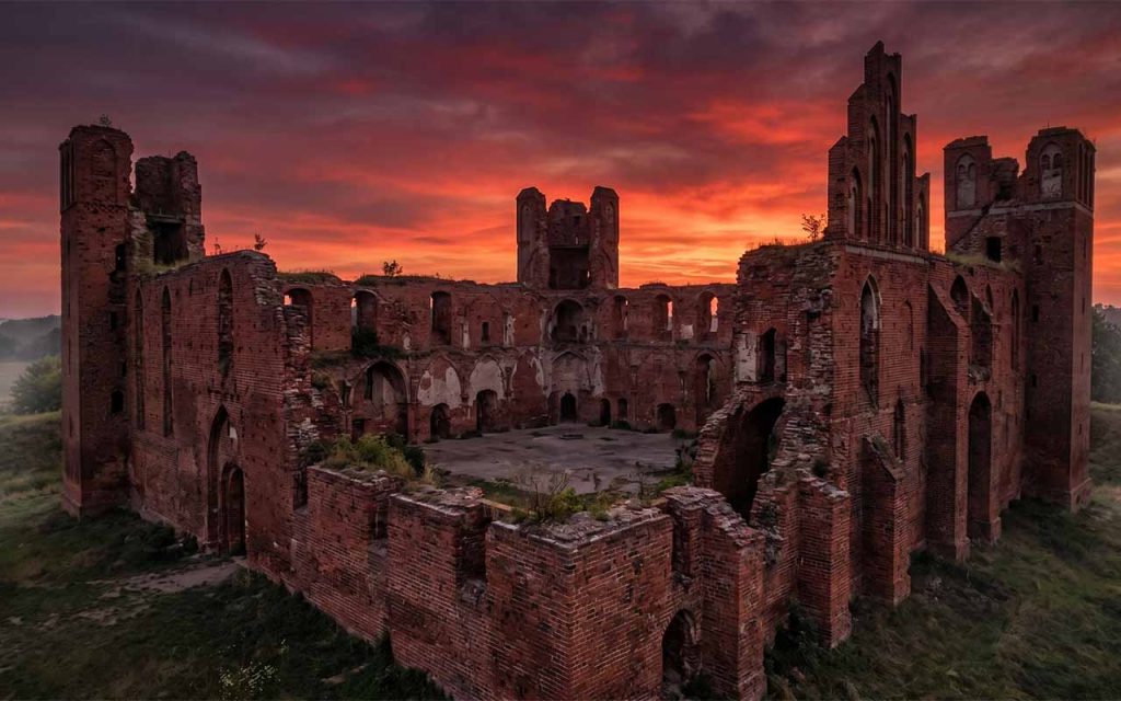 Panorama ruin średniowiecznego zamku krzyżackiego (np. Radzyń Chełmiński), ceglane wieże na tle zachodzącego słońca, dramatyczna atmosfera
